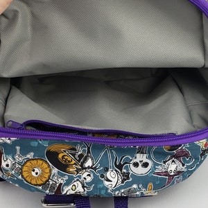 May include: A purple and blue backpack with a zippered pocket and a printed design of characters from the Nightmare Before Christmas. The backpack has a purple strap.