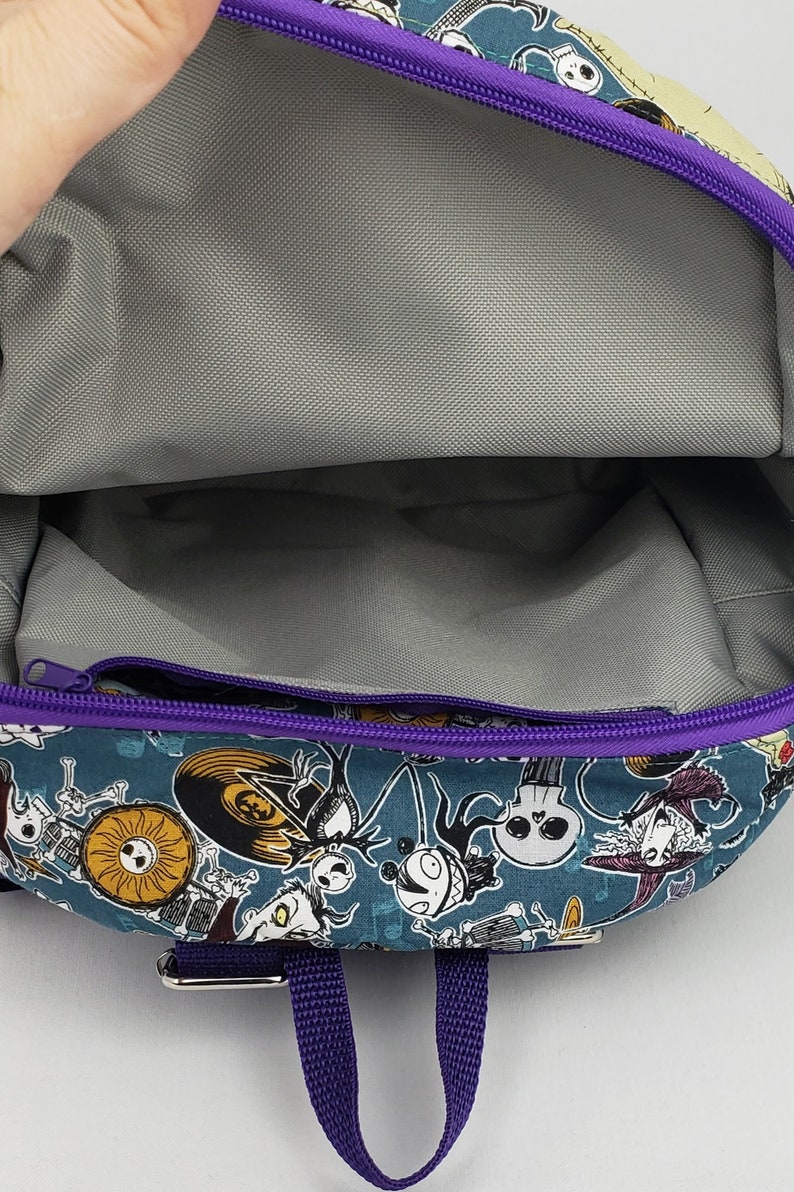 May include: A purple and blue backpack with a zippered pocket and a printed design of characters from the Nightmare Before Christmas. The backpack has a purple strap.