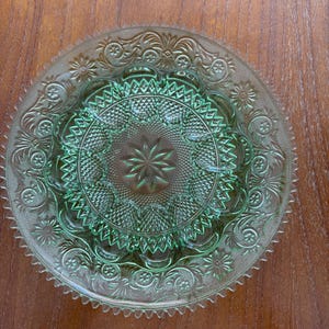 May include: A round, light green pressed glass plate featuring a central starburst pattern surrounded by intricate geometric diamond textures and a scalloped, serrated edge. The glass surface displays elaborate embossed floral and scroll motifs.