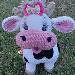 May include: A crocheted plush cow toy with a pink nose, black and white spots, and a pink bow with white flowers. The cow has blue eyes and a bell around its neck.