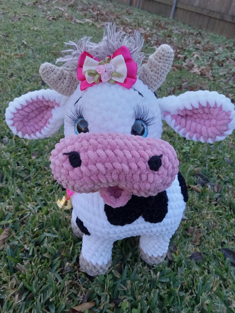 May include: A crocheted plush cow toy with a pink nose, black and white spots, and a pink bow with white flowers. The cow has blue eyes and a bell around its neck.