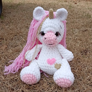 May include: A white crocheted unicorn stuffed animal with pink yarn hair and a gold horn. The unicorn has pink accents on its hooves and a pink heart on its chest.
