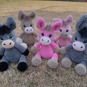 May include: Five handmade plush donkeys in various colors: gray, brown, and pink. Each donkey has large, floppy ears, a white muzzle, and black accents. They are sitting on a bed of dry grass.