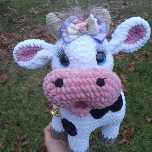 May include: A crocheted plush cow toy with a white body, pink ears and nose, black spots, and a purple bow. The cow has blue eyes and a gold bell around its neck.