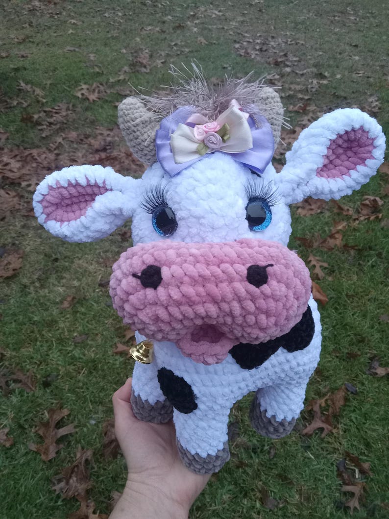May include: A crocheted plush cow toy with a white body, pink ears and nose, black spots, and a purple bow. The cow has blue eyes and a gold bell around its neck.