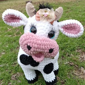May include: A crocheted plush cow toy with white fur, black spots, and a pink nose. The cow has blue eyes with eyelashes and a pink bow with a gold flower on its head.