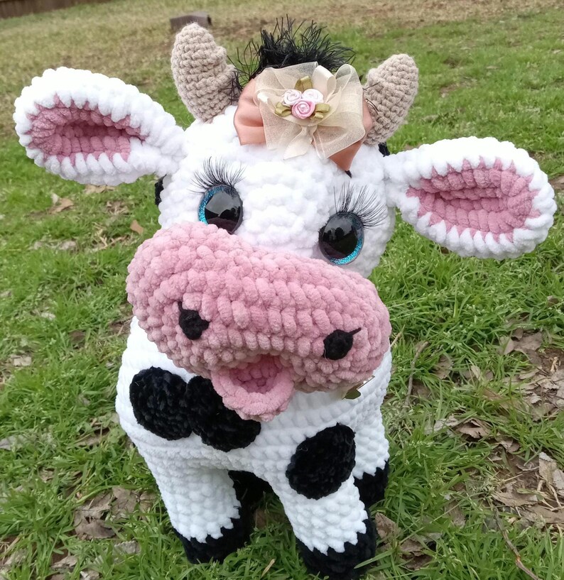May include: A crocheted plush cow toy with white fur, black spots, and a pink nose. The cow has blue eyes with eyelashes and a pink bow with a gold flower on its head.