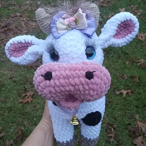 May include: A crocheted plush cow toy with a pink nose, white fur, and black spots. The cow has a purple bow with pink flowers on its head and blue eyes with eyelashes. The cow is holding a gold bell.
