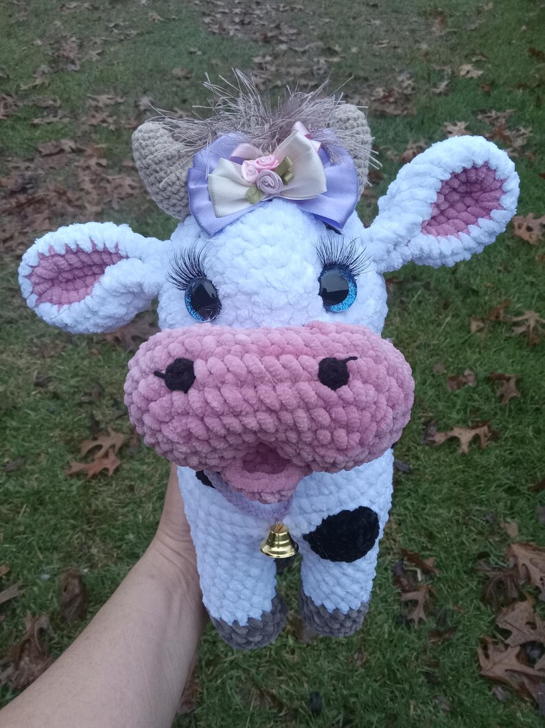 May include: A crocheted plush cow toy with a pink nose, white fur, and black spots. The cow has a purple bow with pink flowers on its head and blue eyes with eyelashes. The cow is holding a gold bell.