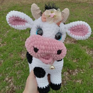 May include: A crocheted plush cow toy with black and white spots, pink ears, and a pink nose. The cow has blue eyes and a pink bow with a gold bell around its neck.