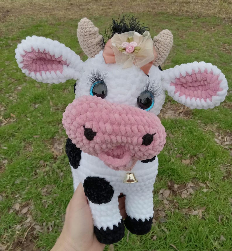 May include: A crocheted plush cow toy with black and white spots, pink ears, and a pink nose. The cow has blue eyes and a pink bow with a gold bell around its neck.