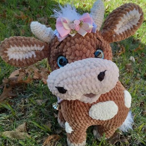 May include: A brown crocheted cow plush toy with white spots, blue eyes, and a pink bow with white flowers. The toy has a bell attached to its neck.