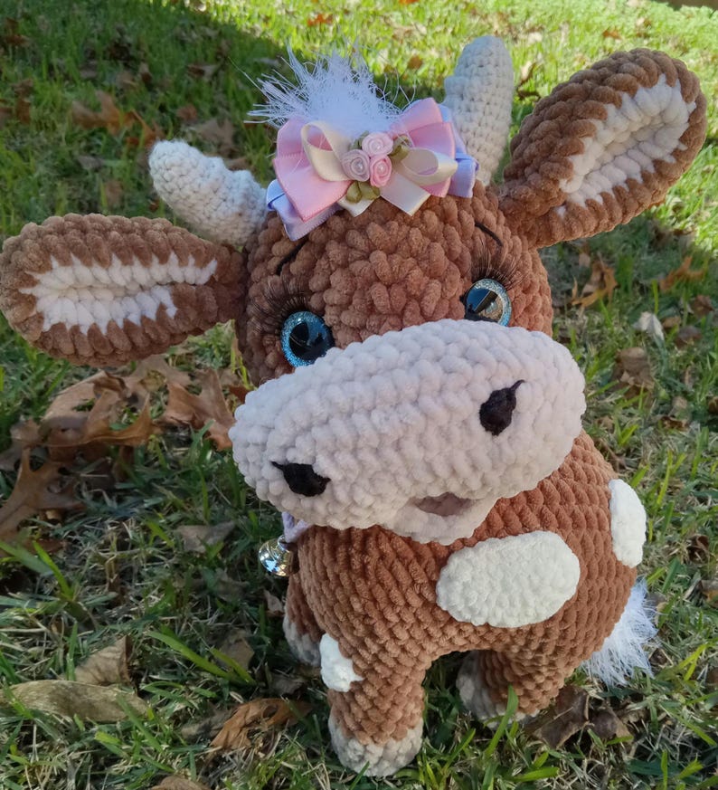 May include: A brown crocheted cow plush toy with white spots, blue eyes, and a pink bow with white flowers. The toy has a bell attached to its neck.