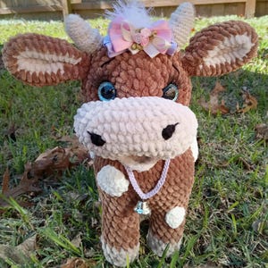 May include: A brown and white crocheted cow plush toy with a pink bow and a bell around its neck. The cow has blue eyes and a white spot on its forehead.