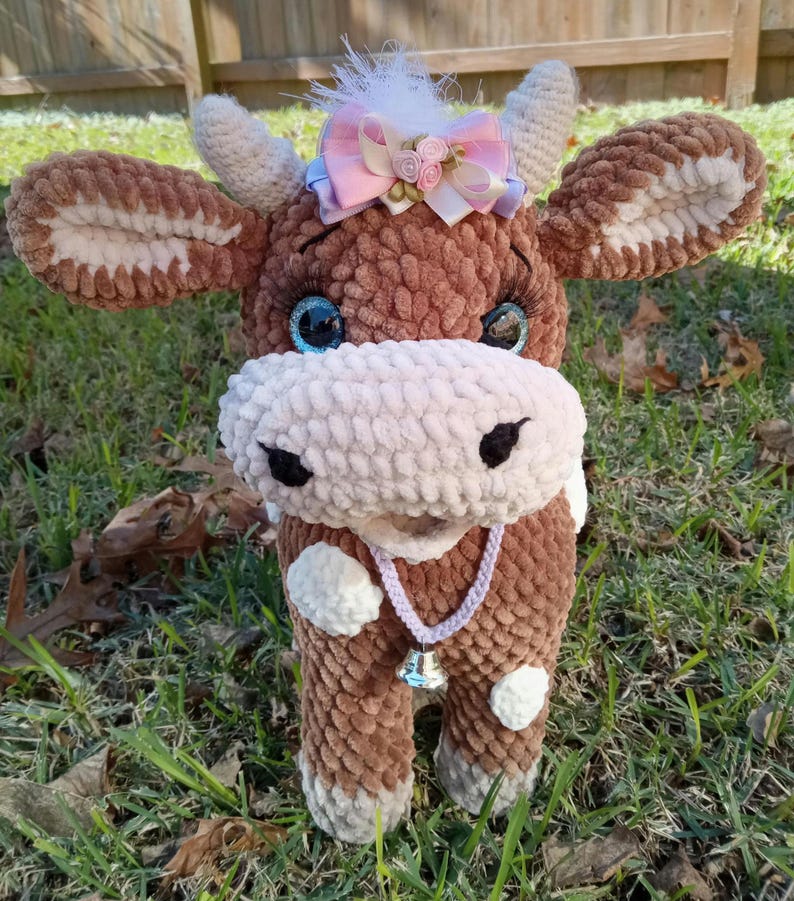 May include: A brown and white crocheted cow plush toy with a pink bow and a bell around its neck. The cow has blue eyes and a white spot on its forehead.