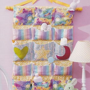Pode incluir: Um organizador de parede de tecido multicolorido com 12 bolsos. Os bolsos são decorados com estrelas, corações e luas. O organizador está pendurado em uma parede com uma fita amarela e uma haste de madeira. Há vários itens nos bolsos, incluindo uma garrafa de loção, uma garrafa de lavagem para bebês, um par de sapatos de bebê e um animal de pelúcia.