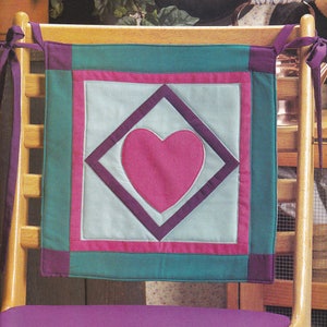 May include: A teal, pink, and purple quilted chair cushion with a heart in the center. The cushion has ties to attach it to a chair.