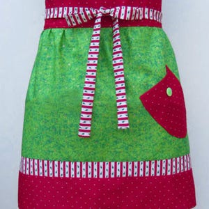 May include: A green and red Christmas-themed apron with a white and red striped tie. The apron has a red pocket with white polka dots and a green button.