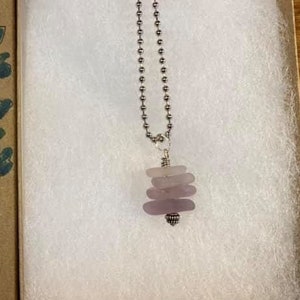 May include: A silver chain necklace with a pendant made of stacked, light purple sea glass. The necklace is displayed in a white gift box with a brown lid. The lid has the text "Kandice Heller Dog Team Designs" written on it.