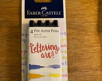 May include: A pack of four Faber-Castell Pitt Artist Pens with India ink. The pens are labeled with the colors blue, yellow, red, and green. The packaging features the text "Lettering art" and "Creative Studio Quality".