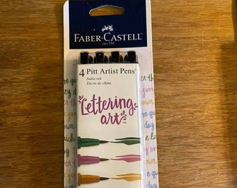 May include: A package of four Faber-Castell Pitt Artist Pens in a variety of colors, including turquoise, pink, gold, and blue. The pens are labeled "Lettering art" and are designed for calligraphy and lettering.