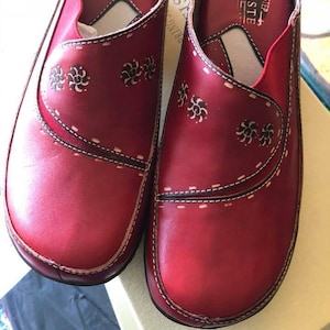 May include: Pair of red leather slip-on shoes with a floral design and the text "Spring Step - L'Artiste" on the insole.
