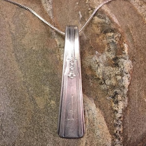 May include: A silver spoon necklace with a decorative design and the letter "I" engraved on the front. The spoon is hanging from a silver chain on a brown textured surface.