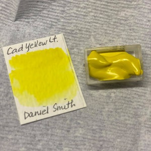 May include: A small container of bright yellow paint labeled "Cad Yellow Lt." with the artist's name "Daniel Smith" written below.