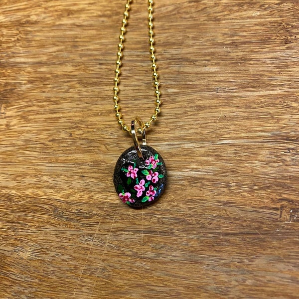 Small oval rock painted with pink tiny flowers and green leaves, glossed, hanging on handmade jumpring, and gold plated ball chain.