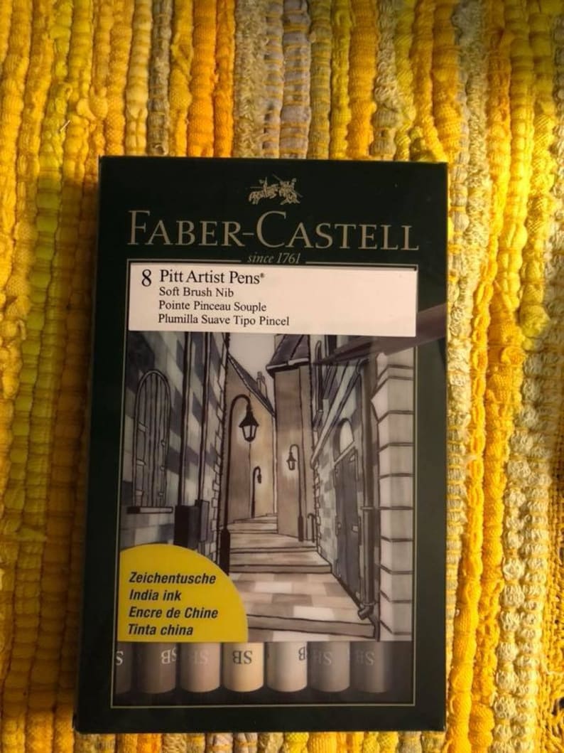 Puede incluir: Una caja verde oscuro de rotuladores Faber-Castell Pitt Artist. La caja presenta una ilustraci&oacute;n de una escena callejera europea. La caja contiene 8 rotuladores de punta de pincel suave con tinta china. El texto de la caja dice "8 Pitt Artist Pens" y "desde 1761".