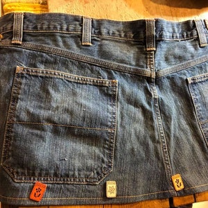 May include: A blue denim skirt with four patches sewn on. The patches are different colors and have various designs. The skirt has two large pockets on the back.