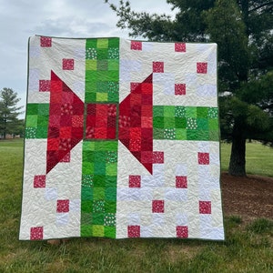 May include: A white, green, and red patchwork quilt with a large red bow in the center. The quilt is made of squares and rectangles of different fabrics.