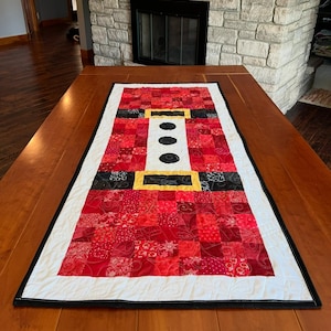 Puede incluir: Camino de mesa con temática de Papá Noel. El camino de mesa presenta un diseño de patchwork en tonos rojos, negros y blancos, con un detalle de hebilla amarilla. Los bordes están rematados con un borde negro, creando un acento temático navideño.