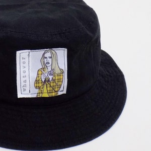 May include: Black bucket hat with a white patch featuring a cartoon image of a woman with blonde hair wearing a yellow and black plaid jacket. The patch also includes the word "whatever" written vertically.