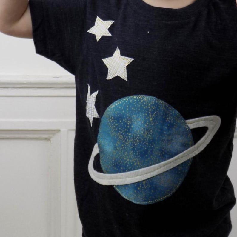 Op de afbeelding: Een donker antracietgrijs T-shirt met korte mouwen en een grote ronde blauwe planeetapplicatie met gouden spikkels en een lichtgrijze ring. Drie witte stervormige applicaties met een subtiel ruitpatroon zijn boven de planeet geplaatst.