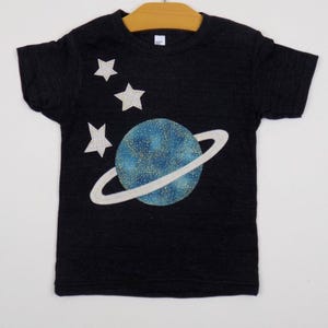 Op de afbeelding: Een zwart t-shirt met korte mouwen voorzien van een afbeelding van een blauwe planeet met een ring en drie witte sterren. De planeet heeft een gespikkelde textuur met goudkleurige stippen. De sterren hebben een ruitjespatroon.