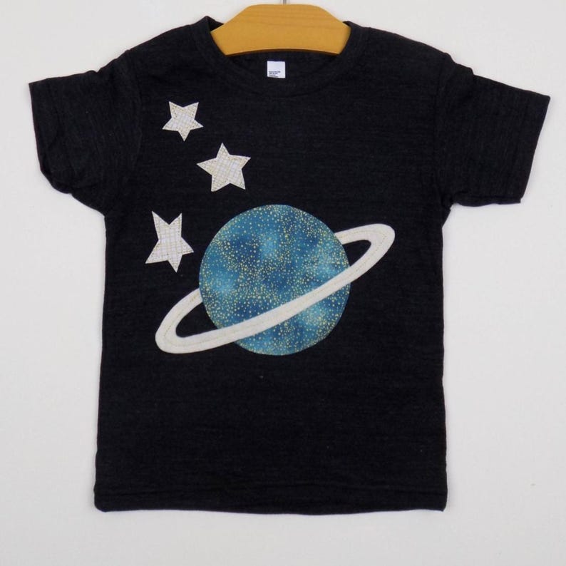 Op de afbeelding: Een zwart t-shirt met korte mouwen voorzien van een afbeelding van een blauwe planeet met een ring en drie witte sterren. De planeet heeft een gespikkelde textuur met goudkleurige stippen. De sterren hebben een ruitjespatroon.