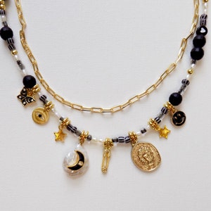 May include: A layered necklace with a gold chain and a beaded strand featuring black and white beads. The necklace has several charms, including a crescent moon, a star, a safety pin, an evil eye, a butterfly, a smiley face, and a coin with a profile of a person.