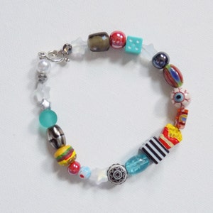 May include: A colorful beaded bracelet with various charms, including a dice, an eye, a hamburger, and a star. The bracelet is made with a mix of glass, metal, and plastic beads.