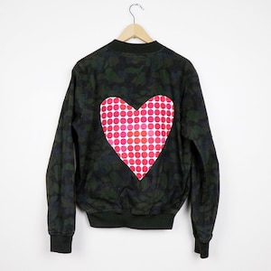May include: A green camouflage bomber jacket with a large red and white polka dot heart on the back.