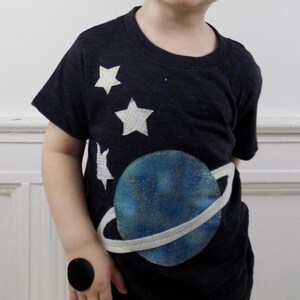 Op de afbeelding: Een donkergrijs t-shirt met korte mouwen en een grote blauwe planeetprint met een witte ring eromheen. Het shirt is versierd met drie witte sterren van verschillende groottes boven de planeet, elk voorzien van een subtiel ruitjespatroon.