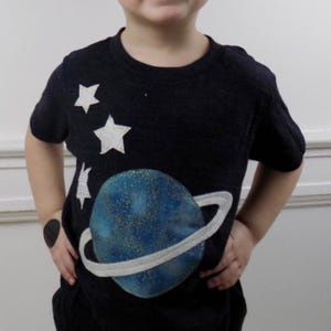 Op de afbeelding: Een donker antracietgrijs t-shirt met korte mouwen en een grote, ronde blauwe planeetafbeelding met gouden spikkels en een lichtgrijze ring. Drie witte, getextureerde stervormige applicaties bevinden zich boven de planeet aan de rechterkant.