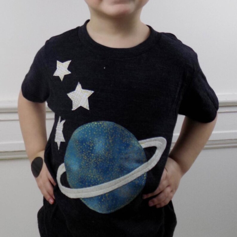 Op de afbeelding: Een donker antracietgrijs t-shirt met korte mouwen en een grote, ronde blauwe planeetafbeelding met gouden spikkels en een lichtgrijze ring. Drie witte, getextureerde stervormige applicaties bevinden zich boven de planeet aan de rechterkant.