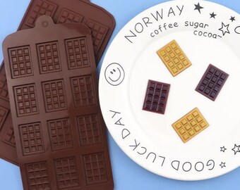 May include: Two brown silicone molds for making chocolate bars. The molds have a waffle pattern. A white plate with the words "Good Luck Day" and "Norway" with the words "coffee, sugar, cocoa" underneath. There are three chocolate bars on the plate, one is brown, one is gold, and one is purple.