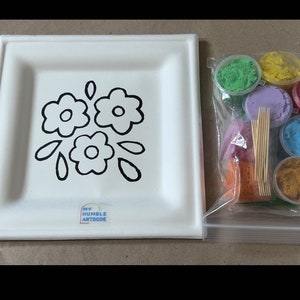 Può includere: Un piatto di carta bianco con un contorno nero di tre fiori e foglie. Il piatto ha un adesivo che dice "My Humble Artbook". Un sacchetto di plastica trasparente contiene sette piccoli contenitori di sabbia colorata, sei spiedini di legno e un piccolo sacchetto di sabbia arancione.