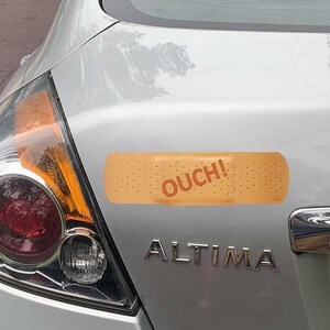 May include: A silver car's rear with a large bandage-shaped sticker. The sticker is tan with small holes and the word "OUCH!" in red. The car's taillight and the word "ALTIMA" are also visible.