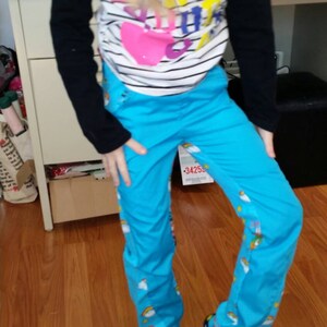 May include: A child wearing a striped long-sleeve shirt with a heart graphic and blue pants with rainbow and cloud patterns. The child is also wearing black slippers with heart and rainbow designs.