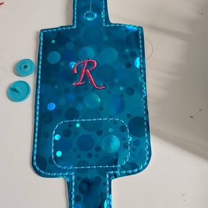 May include: A turquoise blue fabric wristlet with a clear plastic window and a white stitched border. The wristlet has a circle pattern and the letter "R" embroidered in red.
