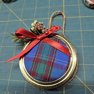 May include: A blue, green and red tartan fabric ornament with a red ribbon and a sprig of faux greenery. The ornament is attached to a twine string.
