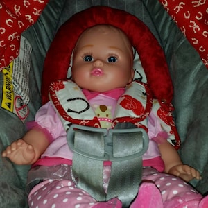 May include: A baby doll wearing a pink and red cowboy-themed outfit with a white polka dot pattern. The doll is sitting in a car seat with a red and white patterned headrest. The doll has blue eyes and pink cheeks.